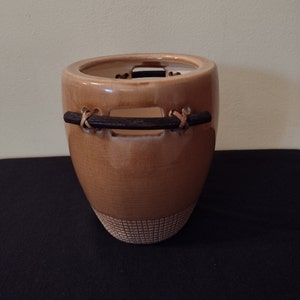 May include: A light brown ceramic planter with a woven leather strap across the opening. The planter has a textured surface and a woven pattern at the bottom.