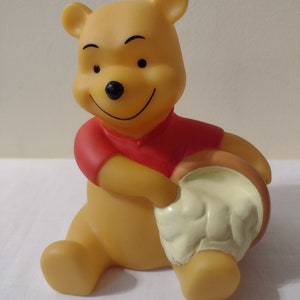 May include: A yellow plastic Winnie the Pooh figurine holding a yellow honey pot. The figurine is sitting down with a red shirt and a smiling face.