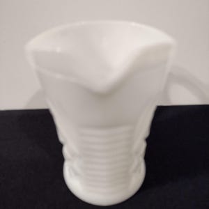May include: A white creamer jug with a spout and handle. The body of the jug has a ribbed design. The jug is made of opaque white glass and is set against a black surface. A classic design.