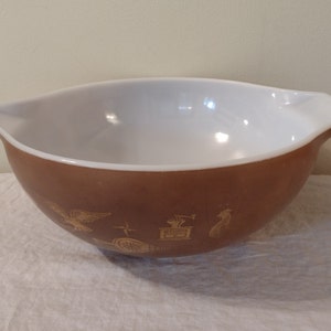 Könnte beinhalten: Eine braune Keramik-Schüssel mit weißem Innenraum und einem goldenen Design mit einem Vogel, einem Stern, einem Rad, einem Gebäude und einem Hahn.