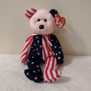 Pode incluir: Um urso de pelúcia Beanie Baby rosa e branco vestindo um padrão de bandeira americana vermelho, branco e azul. O urso tem um nariz preto e olhos pretos. O urso está sentado em uma superfície branca. O urso tem uma etiqueta Ty presa à sua orelha.