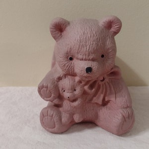 May include: A pink ceramic teddy bear holding a smaller pink teddy bear. The larger bear has a pink bow around its neck.