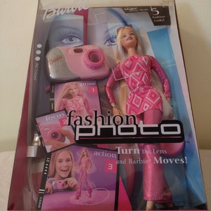 May include: Barbie Fashion Photo doll with a pink and white outfit, long blonde hair, and a pink camera. The box features the text "Make More Than 5 Fashion Looks!" and "action Turn the Lens and Barbie Moves!"