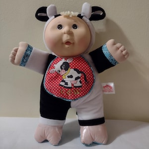 Puede incluir: Una muñeca Cabbage Patch Kid vestida con un disfraz de vaca blanco y negro con un babero rojo que dice "COW". La muñeca tiene el pelo rubio y los ojos marrones.