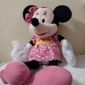 Op de afbeelding: Een grote roze pluche Minnie Mouse met een witte stippenjurk, een gele strik en zwarte schoenen.