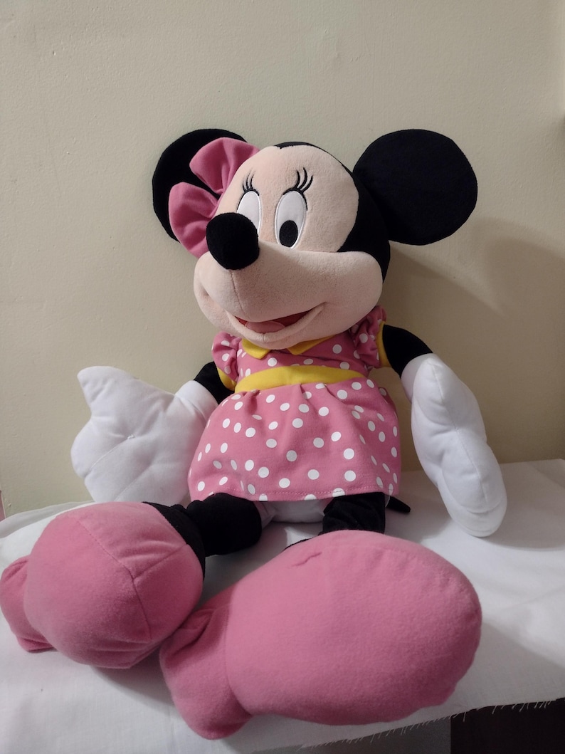 Op de afbeelding: Een grote roze pluche Minnie Mouse met een witte stippenjurk, een gele strik en zwarte schoenen.