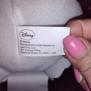 Op de afbeelding: Wit etiket met zwarte tekst die luidt: "Disney &copy;Disney Gemaakt onder licentie van Just Play, LLC 401 Fairway Drive, Deerfield Beach, FL 33441, USA"