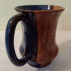 May include: A handmade ceramic mug with a blue and brown glaze. The mug has a unique design with a swirl pattern on the brown side. The handle is dark blue.