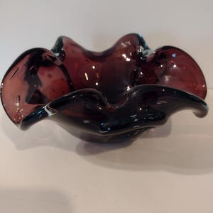 May include: A dark purple glass bowl with a wavy, organic shape. The bowl has a glossy finish and appears to be hand-blown, with slight variations in the glass. The bowl's edges curve outwards, creating a unique, decorative piece.