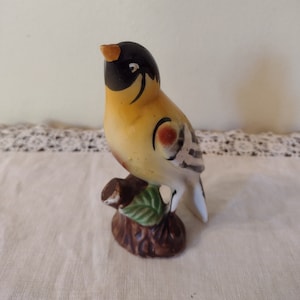 Könnte beinhalten: Eine Keramik-Vogelfigur, bemalt in Schwarz, Weiß, Gelb und Braun. Der Vogel sitzt auf einem braunen Ast mit einem grünen Blatt.