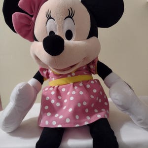 Op de afbeelding: Een groot pluche speelgoed van Minnie Mouse, die een roze gebloemde jurk draagt met een gele riem en een roze strik.