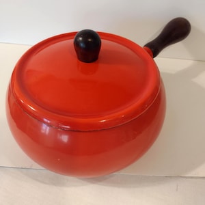 May include: A round, bright red cooking pot with a matching lid and a dark wooden handle. The lid has a dark wooden knob. The pot is likely made of enameled metal. The handle is attached to the side of the pot.