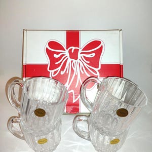 May include: Four clear crystal pitchers with handles, stacked in pairs, are displayed in front of a gift box with a red and white bow design. Each pitcher has a gold sticker. The pitchers are ribbed and have a classic design.