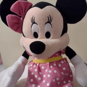 Op de afbeelding: Een pluche speelgoed van Minnie Mouse die een roze jurk met witte stippen draagt met een gele riem en een roze strik. Ze heeft grote zwarte ogen en een zwarte neus.