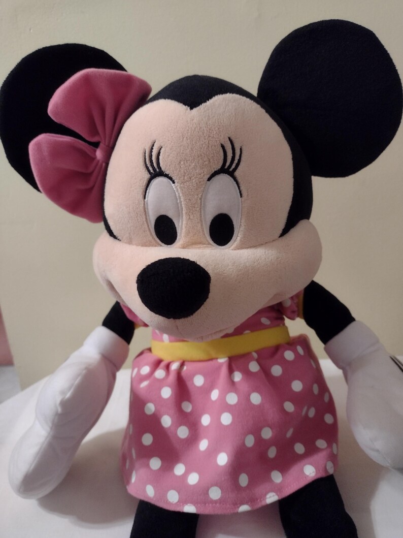 Op de afbeelding: Een pluche speelgoed van Minnie Mouse die een roze jurk met witte stippen draagt met een gele riem en een roze strik. Ze heeft grote zwarte ogen en een zwarte neus.