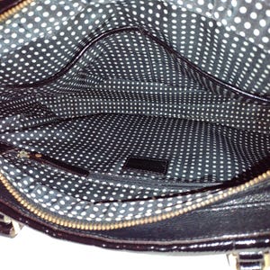 Op de afbeelding: De binnenkant van een zwarte laklederen handtas met een witte stippenvoering. De tas heeft een ritssluiting en een klein binnenvak.