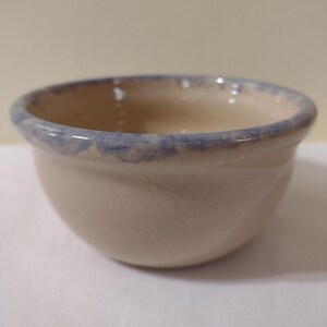 May include: A small, beige ceramic bowl with a light blue rim. The bowl is slightly rounded and has a smooth, glazed finish.