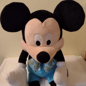 Może przedstawiać: Pluszowa zabawka Mickey Mouse ubrana w niebieską koszulę z żółto-niebieskim wzorem. Zabawka ma duże czarne uszy, białe rękawiczki i żółte buty.