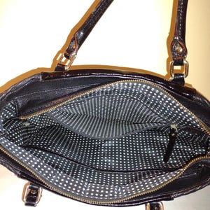 Op de afbeelding: Zwarte leren tote bag met een witte stippenvoering. De tas heeft twee bovenste handvatten en een ritssluiting.