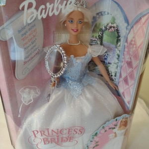 Puede incluir: Muñeca Barbie Princesa Novia con un vestido blanco y azul, una tiara plateada y un espejo de mano plateado. La muñeca sostiene el espejo y lo mira. La caja tiene un fondo rosa con un castillo y flores. El texto de la caja dice "Barbie Princesa Novia".
