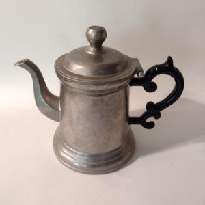 May include: A small, antique pewter teapot with a black, ornate handle. The teapot has a curved spout and a lid with a small knob. The metal has a slightly aged appearance, suggesting it is vintage.