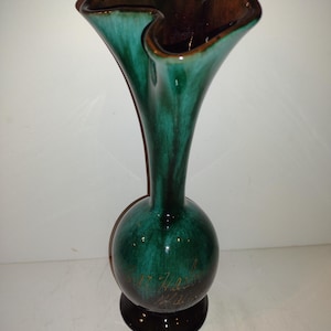 May include: A tall, ceramic vase with a flared, ruffled rim. The vase features a dark brown interior and a glossy, teal green exterior. The base is a dark brown color. The vase has a bulbous midsection and a slender neck.
