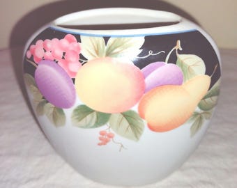May include: A white ceramic vase with a black rim and a fruit design. The vase features a yellow orange, a purple plum, a yellow pear, and a bunch of pink grapes.
