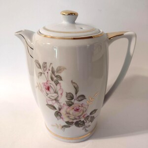 May include: White ceramic coffee pot with a lid and handle. The pot is decorated with pink roses and green leaves. Gold trim accents the lid, handle, and base. The spout is on the left side.