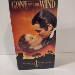 Puede incluir: Una cinta VHS vintage de la película "Lo que el viento se llevó". La portada presenta una imagen romántica de dos personas abrazándose, sobre un fondo de puesta de sol. El título se muestra de forma destacada, junto con el texto de los premios Oscar.