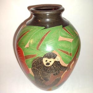 Może przedstawiać: Ręcznie malowany ceramiczny wazon z ciemnobrązową obwódką i zaokrąglonym korpusem. Wazon ma zielone tło z brązowymi i czerwonymi akcentami oraz szczegółową ilustrację małpy. Małpa jest czarna i brązowa, siedzi na gałęzi.