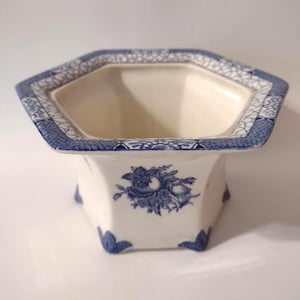 May include: A white and blue ceramic planter with a hexagonal top and a floral design. The planter has a blue patterned border and blue floral accents. The planter is elevated on small feet.