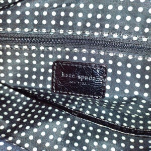 Op de afbeelding: Close-up van de zwarte stoffen voering van een handtas met een wit stippenpatroon. Een zwart lederen label met de tekst "kate spade new york" is zichtbaar.