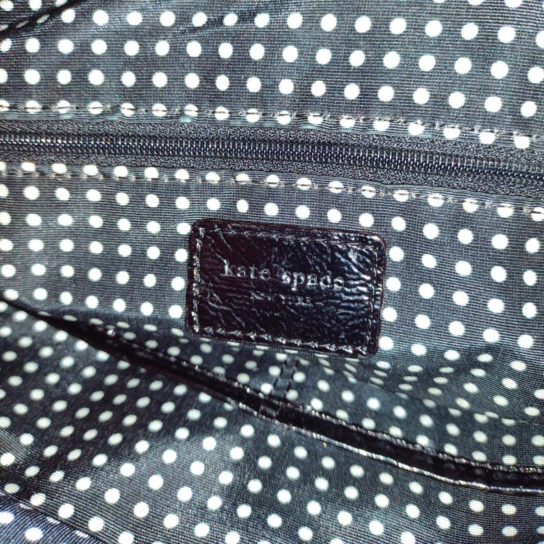 Op de afbeelding: Close-up van de zwarte stoffen voering van een handtas met een wit stippenpatroon. Een zwart lederen label met de tekst "kate spade new york" is zichtbaar.