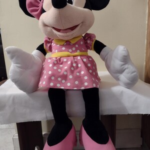Op de afbeelding: Een groot pluche Minnie Mouse speelgoed dat een roze jurk draagt met witte stippen, een gele riem en een roze strik. Ze heeft zwarte schoenen en witte handschoenen.