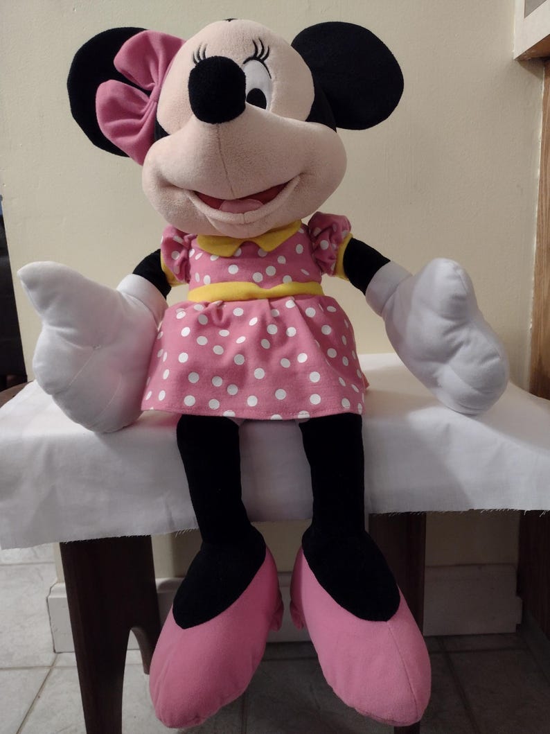 Op de afbeelding: Een groot pluche Minnie Mouse speelgoed dat een roze jurk draagt met witte stippen, een gele riem en een roze strik. Ze heeft zwarte schoenen en witte handschoenen.