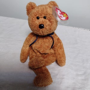 Könnte beinhalten: Ein brauner Teddybär mit einem blauen Band um den Hals. Der Bär sitzt auf einer weißen Oberfläche und hat ein rotes und weißes Ty-Etikett am Ohr.