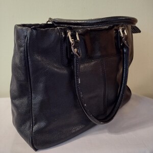 Op de afbeelding: Een zwarte leren tote bag met twee bovenste handvatten. De tas heeft een klep sluiting en een zilveren gesp.