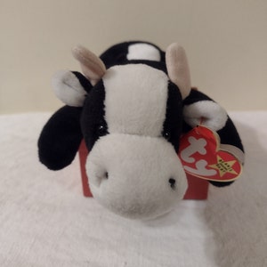 Pode incluir: Vaca de pelúcia preta e branca com uma etiqueta vermelha Ty Beanie Baby. A vaca está deitada com a cabeça voltada para a câmera.
