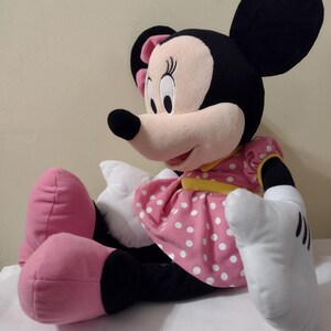 Op de afbeelding: Een groot pluche Minnie Mouse speelgoed dat een roze stippenjurk draagt met een gele band. Ze heeft zwarte oren, een zwarte neus en roze schoenen.