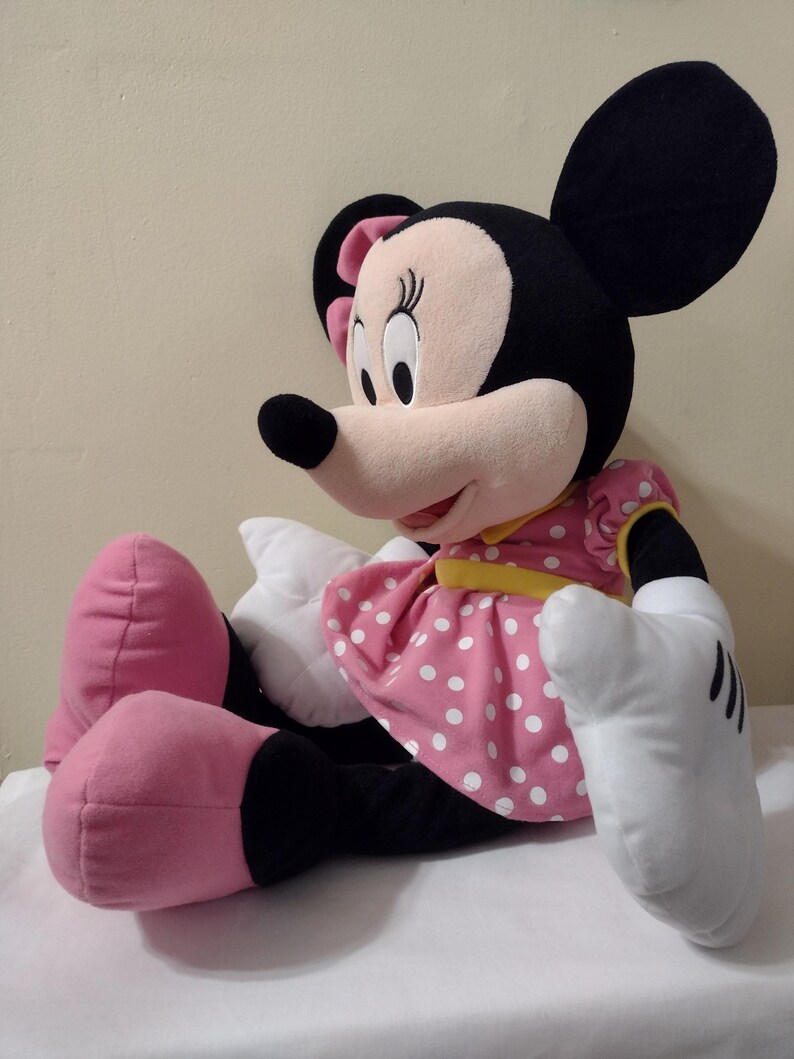 Op de afbeelding: Een groot pluche Minnie Mouse speelgoed dat een roze stippenjurk draagt met een gele band. Ze heeft zwarte oren, een zwarte neus en roze schoenen.