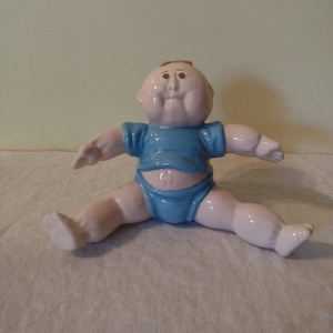 Könnte beinhalten: Eine Keramikfigur eines Babys mit einem blauen Shirt und einer Windel, das mit gespreizten Beinen sitzt. Das Baby hat einen großen Kopf und ein breites Lächeln.