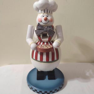 May include: A white wooden nutcracker figurine of a chef wearing a white hat and a red and white striped apron. The chef is holding a pie in his hands. The nutcracker is standing on a blue and white base.