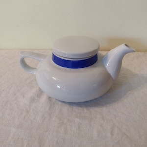 Puede incluir: Tetera de cerámica blanca con una banda azul alrededor de la tapa y el cuerpo. La tetera tiene un cuerpo redondo y un pico en el lado derecho.