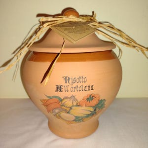 Può includere: Un vaso in terracotta con coperchio e un fiocco di paglia. Il vaso ha un disegno stampato di verdure e il testo "Risotto all'ortolana".