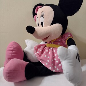 Op de afbeelding: Een groot pluche Minnie Mouse speelgoed zit met gekruiste benen. Ze draagt een roze jurk met witte stippen en een gele sjerp. Haar oren zijn zwart en haar schoenen zijn roze.