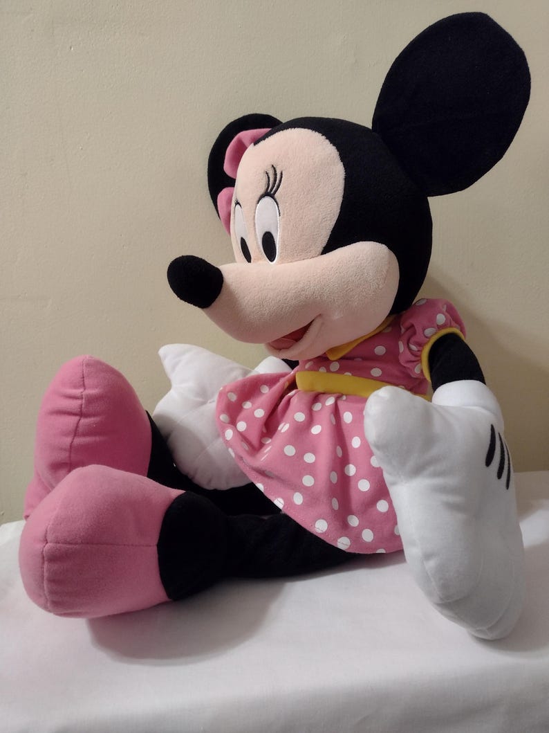 Op de afbeelding: Een groot pluche Minnie Mouse speelgoed zit met gekruiste benen. Ze draagt een roze jurk met witte stippen en een gele sjerp. Haar oren zijn zwart en haar schoenen zijn roze.