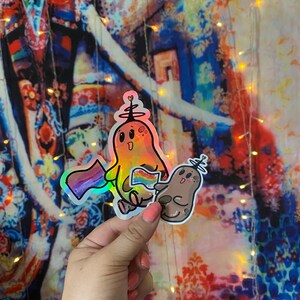 May include: Two iridescent holographic stickers of cartoon characters holding rainbow pride flags. The characters are a yellow and orange creature with a crown and a brown creature with a crown.