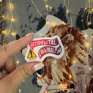 May include: A white sticker with a red and purple gradient design. The sticker has a yellow caution sign with an exclamation point and the text "CAUTION!" The sticker also has a red and purple banner with the text "ACCIDENTAL CANNIBALISM".