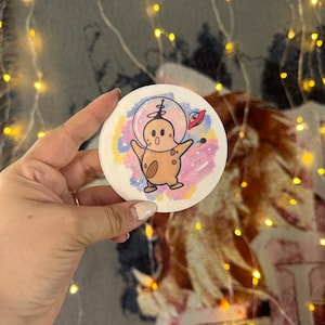 May include: A round sticker with a cartoon character in a space suit. The character is brown with a white face and is wearing a white space suit with a red helmet. The background is a watercolor-style galaxy with pink, blue, and purple colors.