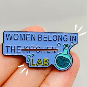 May include: A blue enamel pin with the text "Women belong in the kitchen" crossed out and replaced with "lab". A small cartoon beaker with blue liquid is next to the text.
